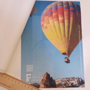 May include: A colorful hot air balloon with the registration number TC-BIV, flying over a rocky landscape. The balloon is against a blue sky with white clouds. The image includes the text "LIGHTWEIGHT WATER RESISTANT 100% POLYESTER-FACE SIDE WATER RESISTANT 105GSM" and the text "the fabric centre".