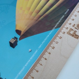 May include: A close-up of a wooden ruler with a hot air balloon printed on a blue background. The ruler is marked with numbers from 5 to 18. The text "LIGHTWEIGHT WATER RESISTANT. 100% POLYESTER-FACE SIDE WATER RESISTANT - 105GSM" is printed on the ruler.