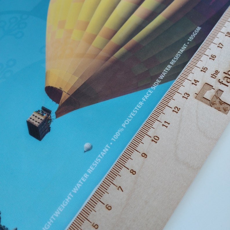 May include: A close-up of a wooden ruler with a hot air balloon printed on a blue background. The ruler is marked with numbers from 5 to 18. The text "LIGHTWEIGHT WATER RESISTANT. 100% POLYESTER-FACE SIDE WATER RESISTANT - 105GSM" is printed on the ruler.