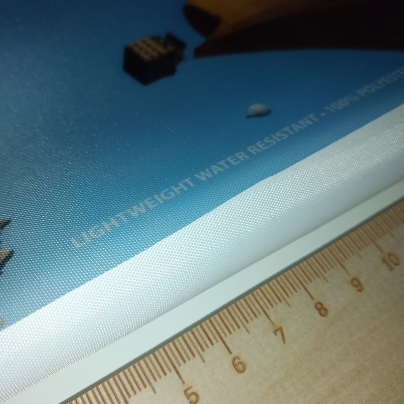 May include: Close-up of a white fabric with the text "Lightweight Water Resistant - 100% Polyester" printed on a blue background. The fabric is being measured with a ruler.