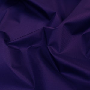 May include: A close-up of a deep purple fabric with a smooth, slightly textured surface. The fabric is folded and draped, creating soft folds and curves.