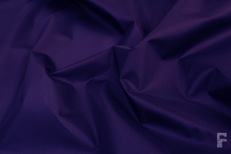 May include: A close-up of a deep purple fabric with a smooth, slightly textured surface. The fabric is folded and draped, creating soft folds and curves.