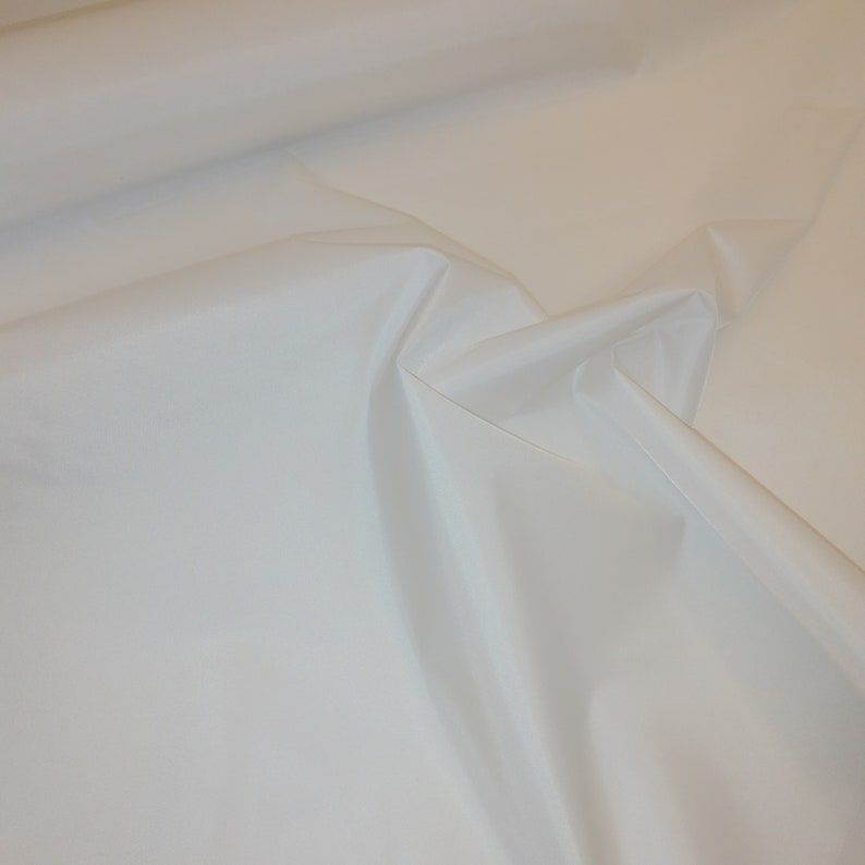 May include: White fabric with a smooth, silky texture. The fabric is folded and draped, showing its soft, flowing quality.