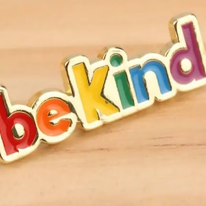 May include: A gold-colored enamel pin with the words "be kind" in rainbow colors. The pin is on a light brown wooden surface. The letters are red, orange, yellow, green, blue, and purple.