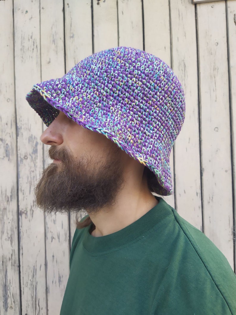 May include: A purple, green, and white crocheted bucket hat. The hat is worn by a person with a beard.