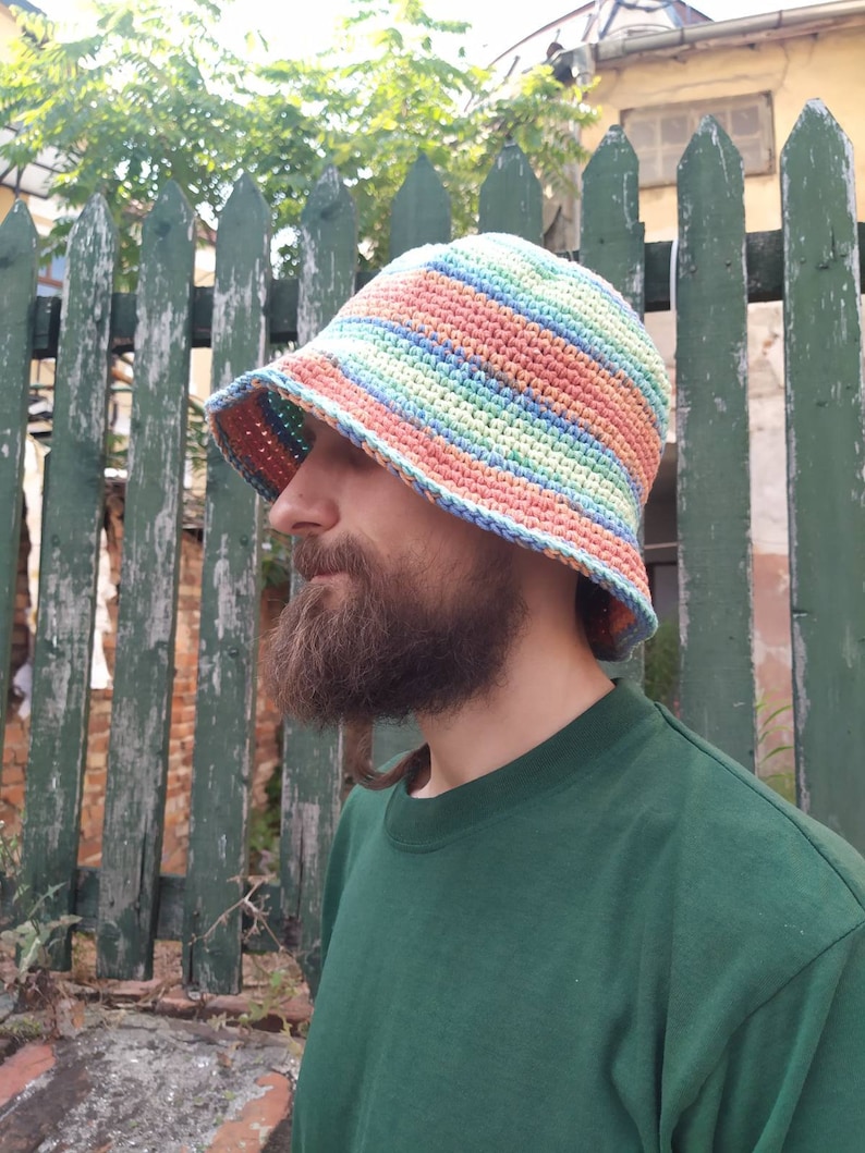 May include: A person wearing a green shirt and a crocheted bucket hat with stripes of pink, blue, and green.