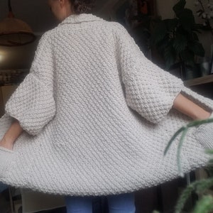 May include: Cream-colored, oversized knit cardigan with a textured stitch pattern. The cardigan features wide sleeves and pockets. The garment is worn open, showcasing its relaxed fit and cozy design. A stylish and comfortable addition to any wardrobe.