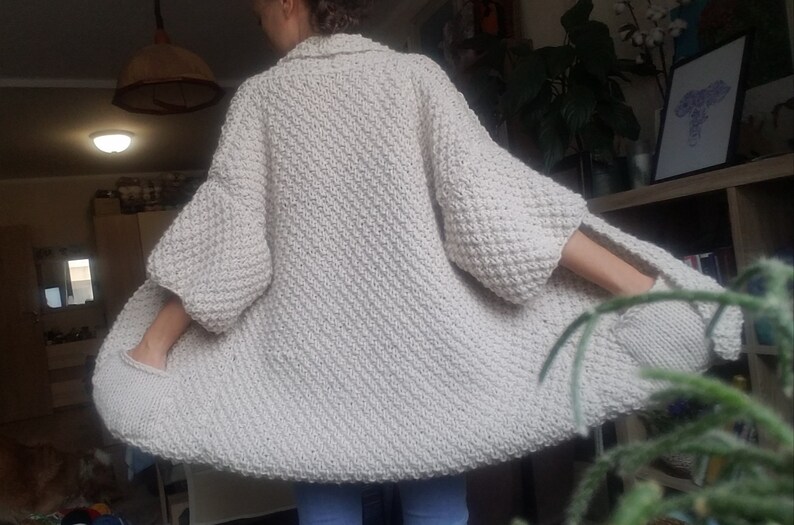 May include: Cream-colored, oversized knit cardigan with a textured stitch pattern. The cardigan features wide sleeves and pockets. The garment is worn open, showcasing its relaxed fit and cozy design. A stylish and comfortable addition to any wardrobe.