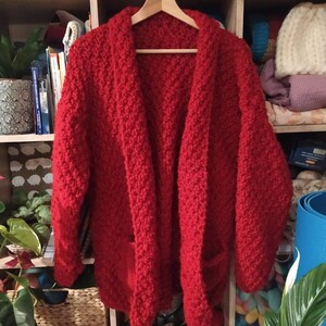 May include: A vibrant red, chunky-knit cardigan with two front pockets hangs on a wooden hanger. The open-front design and textured knit suggest a cozy, handmade garment. The cardigan is displayed against a backdrop of a wooden shelf filled with books and yarn.