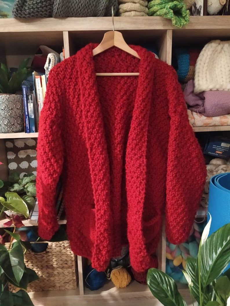May include: A vibrant red, chunky-knit cardigan with two front pockets hangs on a wooden hanger. The open-front design and textured knit suggest a cozy, handmade garment. The cardigan is displayed against a backdrop of a wooden shelf filled with books and yarn.