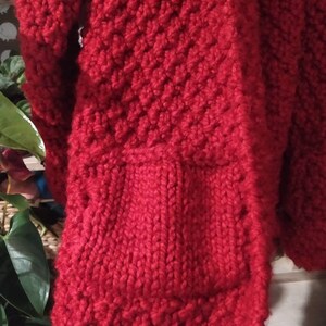 May include: Close-up of a vibrant red knitted garment, possibly a sweater or cardigan. The thick yarn creates a textured, patterned surface with visible stitches. The design includes a pocket. The overall aesthetic suggests a handmade, cozy item.