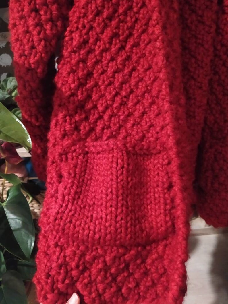 May include: Close-up of a vibrant red knitted garment, possibly a sweater or cardigan. The thick yarn creates a textured, patterned surface with visible stitches. The design includes a pocket. The overall aesthetic suggests a handmade, cozy item.