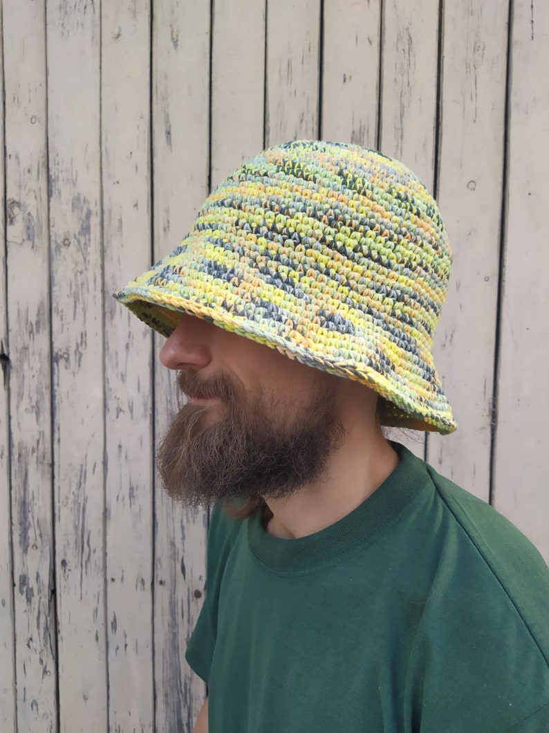 May include: A crocheted bucket hat with a yellow, green, and gray striped pattern. The hat is worn by a person with a beard.
