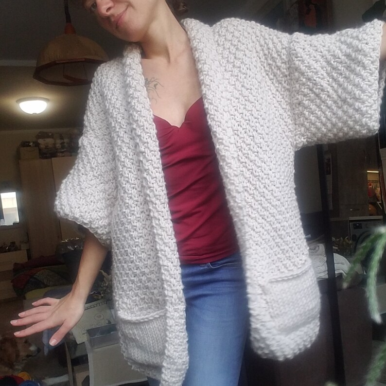 May include: A cream-colored, chunky knit cardigan with short sleeves and two front pockets. The open-front sweater is made of thick yarn and has a textured stitch pattern. The cardigan is worn over a red top and blue jeans.