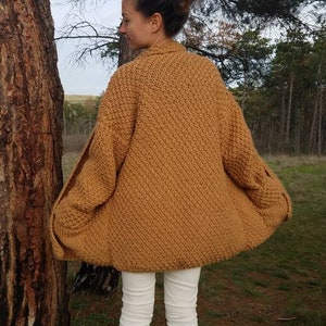 May include: A mustard-colored, knitted cardigan with a textured pattern. The cardigan has a wide, open design and long sleeves. The person is wearing white pants and brown boots. The background includes trees and a natural setting.
