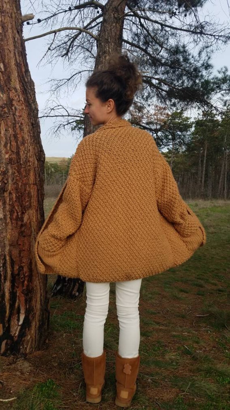 May include: A mustard-colored, knitted cardigan with a textured pattern. The cardigan has a wide, open design and long sleeves. The person is wearing white pants and brown boots. The background includes trees and a natural setting.