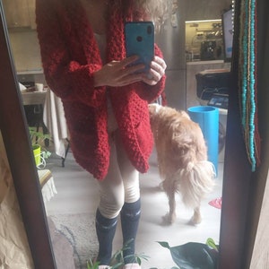 May include: A red, open-front cardigan with a textured knit design. The long-sleeved sweater has two front pockets. The person in the image is wearing a white top, light-colored leggings, and dark socks. A golden retriever dog is in the background.