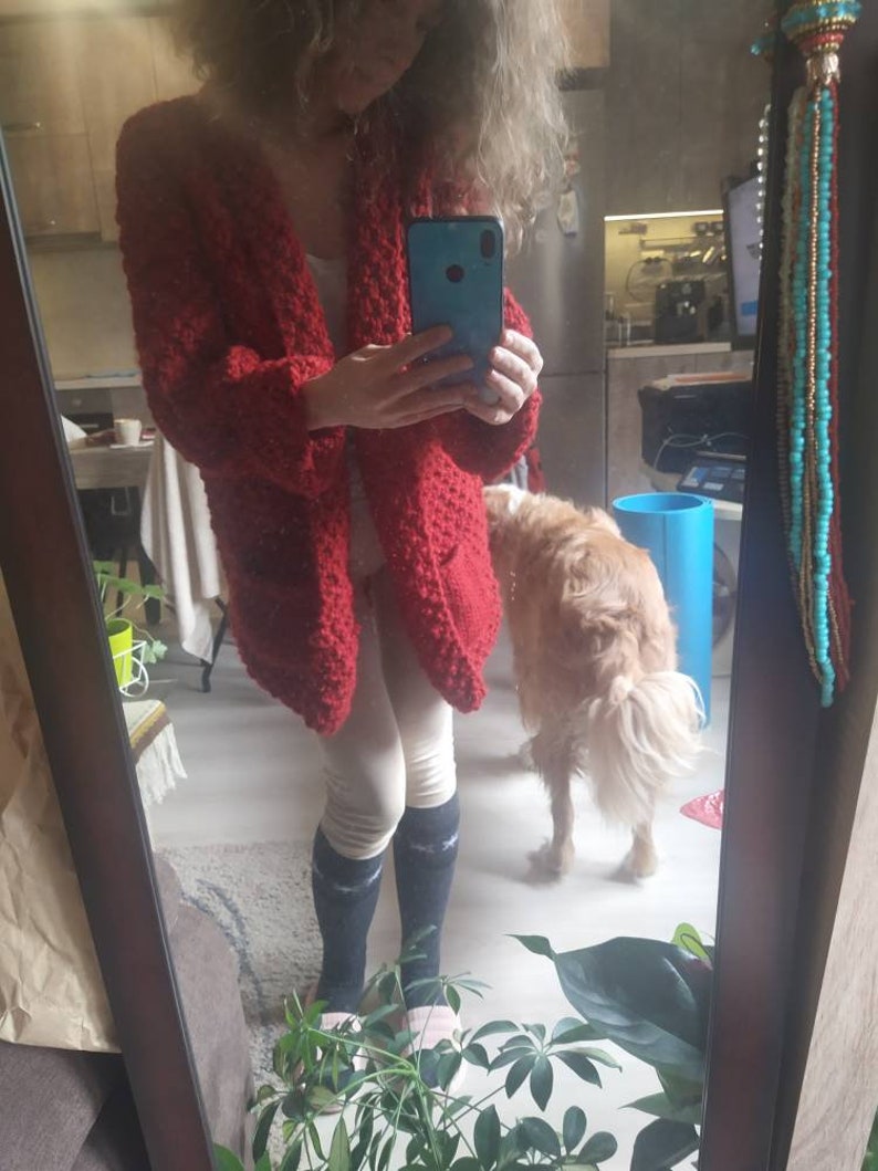 May include: A red, open-front cardigan with a textured knit design. The long-sleeved sweater has two front pockets. The person in the image is wearing a white top, light-colored leggings, and dark socks. A golden retriever dog is in the background.