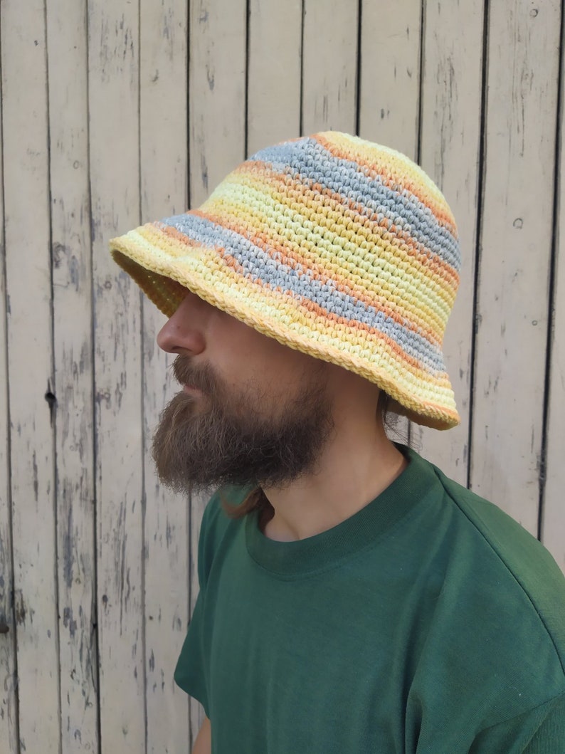 May include: A yellow, orange, and gray striped crocheted bucket hat. The hat is worn by a person with a beard.