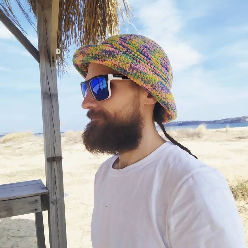 May include: A person wearing a colorful crocheted bucket hat with blue sunglasses and a white t-shirt. The person has a long braid and a beard.
