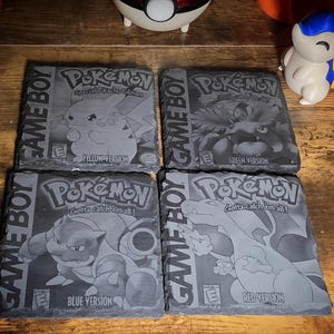 May include: Four slate coasters featuring Pokémon game covers. Each coaster displays a different version: Yellow, Green, Blue, and Red. The coasters have the Game Boy logo and the Pokémon logo. The coasters are gray with white lettering and images.