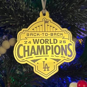 May include: A gold-colored Christmas tree ornament with the text "BACK-TO-BACK 24 WORLD 25 CHAMPIONS" and the LA logo. The ornament is hanging on a green Christmas tree with white and red ornaments.