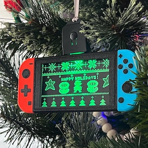 May include: A black and blue Nintendo Switch Christmas ornament with a green pixelated screen displaying "Happy Holidays!" and a pixelated snowman. The ornament has a black and white cord for hanging.