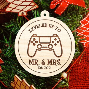 May include: A round wooden Christmas ornament with the text "LEVELED UP TO MR. & MRS. Est. 2021" and a controller graphic. The ornament is hanging on a Christmas tree with red and white decorations.
