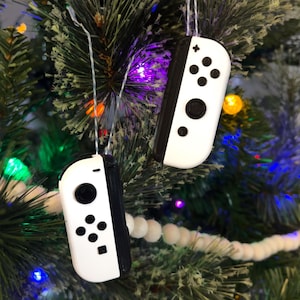 May include: Two white and black Nintendo Switch Joy-Con controllers are hanging from a Christmas tree as ornaments. The controllers are decorated with the standard buttons and a plus sign on the top.