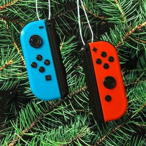 May include: Two Nintendo Switch Joy-Con controllers, one blue and one red, hanging from a Christmas tree. The controllers are decorated with black buttons and a black border.