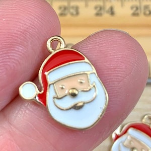 May include: A gold-colored Santa Claus charm with a red hat, white beard, and a smiling face. The charm is made of enamel and is likely used for jewelry making or crafting. The charm is held in a person's hand.
