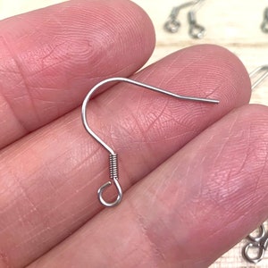May include: Close-up of several silver-colored earring hooks. The hooks are made of thin metal wire, with a curved hook at one end and a small loop and coil at the other. The hooks are on a light-colored wooden surface.