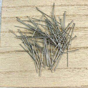 May include: A collection of silver-colored straight pins scattered on a light-colored wooden surface. The pins have small, flat heads and are of varying lengths. The pins are likely used for sewing or crafting projects.