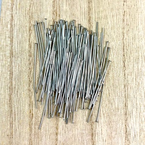May include: A collection of silver-colored head pins scattered on a light brown wooden surface. The pins have a small, flat head and a long, slender body. The pins are used for crafting and sewing projects.