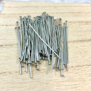 May include: A pile of silver-colored head pins with flat, round heads and long, thin shafts. The pins are scattered on a light-colored wooden surface, creating a textured and organized appearance. These pins are likely used for crafting or sewing projects.