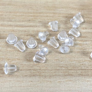 May include: A collection of clear, translucent earring backs scattered on a light wood surface. The earring backs are small, with a rounded base and a tapered, cylindrical post hole. They appear to be made of a soft, flexible material.