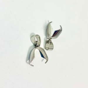May include: Two silver-colored pendant bails, designed for jewelry making. Each bail features a curved, open-ended clasp and a small loop for attaching to a chain or cord. The bails are made of a shiny, metallic material.
