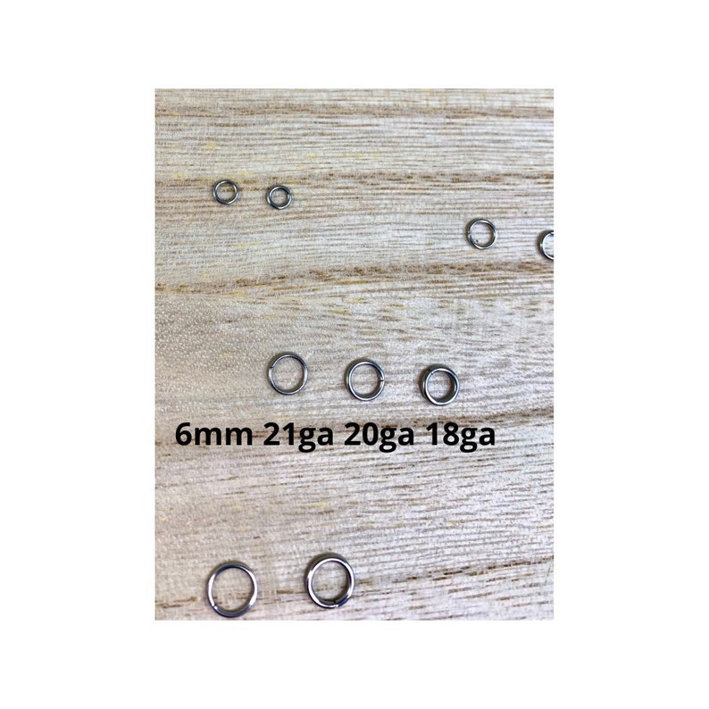May include: A collection of small, silver-colored metal rings of varying sizes, arranged on a light wood surface. The text "6mm 21ga 20ga 18ga" is printed below some of the rings. These rings are likely used for jewelry making or crafts.