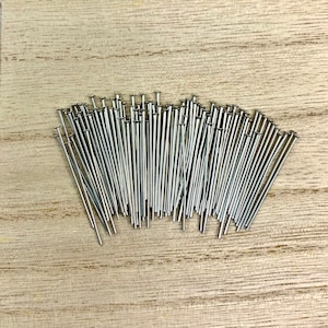 May include: A collection of silver-colored straight pins with small, flat heads. The pins are arranged in a loose cluster on a light-colored wooden surface, creating a contrast between the shiny metal and the wood grain.
