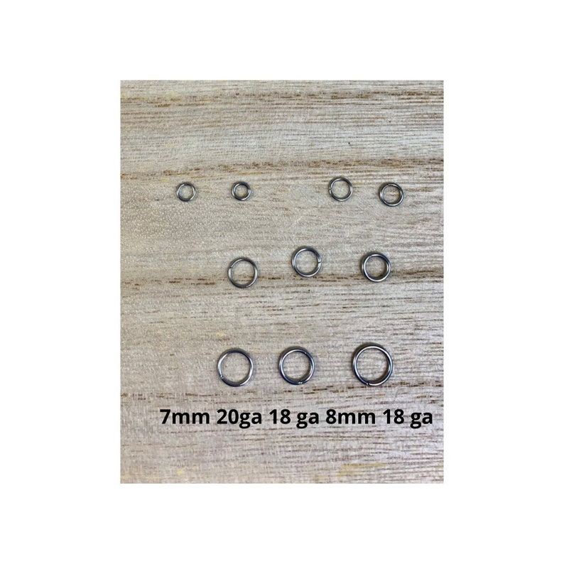 May include: A collection of small, silver-colored jump rings of varying sizes, arranged on a light wood surface. The text "7mm 20ga 18 ga 8mm 18 ga" is printed at the bottom. These rings are likely used for jewelry making or crafts.