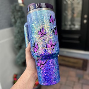 Może przedstawiać: Błyszczący kubek podróżny z fioletową pokrywką i uchwytem. Kubek pokryty jest niebieskim i fioletowym brokatem i ozdobiony kilkoma fioletowymi naklejkami z motylami. Zaprojektowany do utrzymywania gorących lub zimnych napojów.