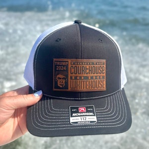 Op de afbeelding: Een zwarte en witte truckercap met een bruin leren patch waarop staat "Trump 2024 From the Courthouse to the Whitehouse". De pet is gemaakt door Richardson en heeft maat 112.