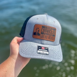 May include: A gray and black trucker hat with a brown leather patch that says "Make Convicted Felons Great Again". The patch features a picture of a man. The hat is made by Richardson and is adjustable.