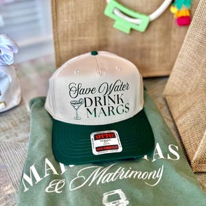 May include: A beige and dark green baseball cap with the text Save Water DRINK MARGS and a margarita glass graphic on the front. The cap features an Otto brand sticker on the brim. It is placed on a green t-shirt with white text and graphics.
