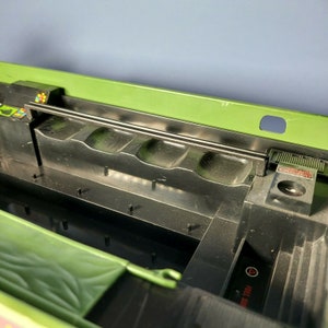 May include: A green plastic toy vehicle with a black interior. The interior features a black plastic bar and four moulded seats. The vehicle has a green plastic canopy and a black plastic handle. The vehicle has a red button labelled "Fuel Tank".