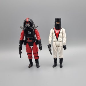 May include: Two action figures, one in a black and red suit with a skull and crossbones on the chest, and the other in a white suit with a red skull and crossbones on the chest.
