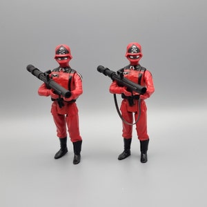 Könnte beinhalten: Zwei rote Actionfiguren mit schwarzen Helmen und Totenkopf-Abzeichen, die schwarze Waffen halten. Die Figuren tragen rote Hosen und schwarze Stiefel.