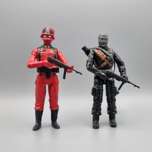 Könnte beinhalten: Zwei Actionfiguren, eine in Rot mit einem schwarzen Totenkopf-Helm und die andere in Schwarz mit einer grauen Gesichtsmaske, beide halten Waffen.