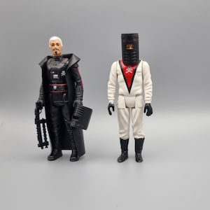 Puede incluir: Dos figuras de acción, una con un atuendo negro y rojo con una capa y un bláster, y la otra con un atuendo blanco con un casco negro y un símbolo rojo de calavera y huesos cruzados en el pecho.