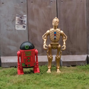 May include: Two action figures: a gold-colored skeletal robot and a red and black droid. The robot has a skull-like face and a blue and red panel on its chest. The droid has a black dome and green and red details. They stand on green grass.